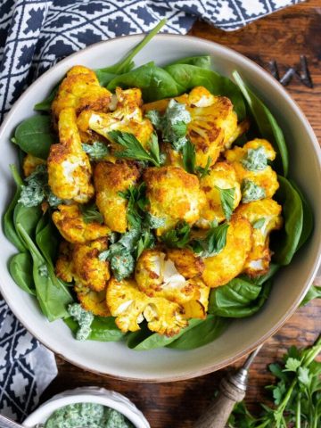 A bowl of bright yellow turmeric roasted cauliflower on a bed of spinach topped with a fresh herb dressing.