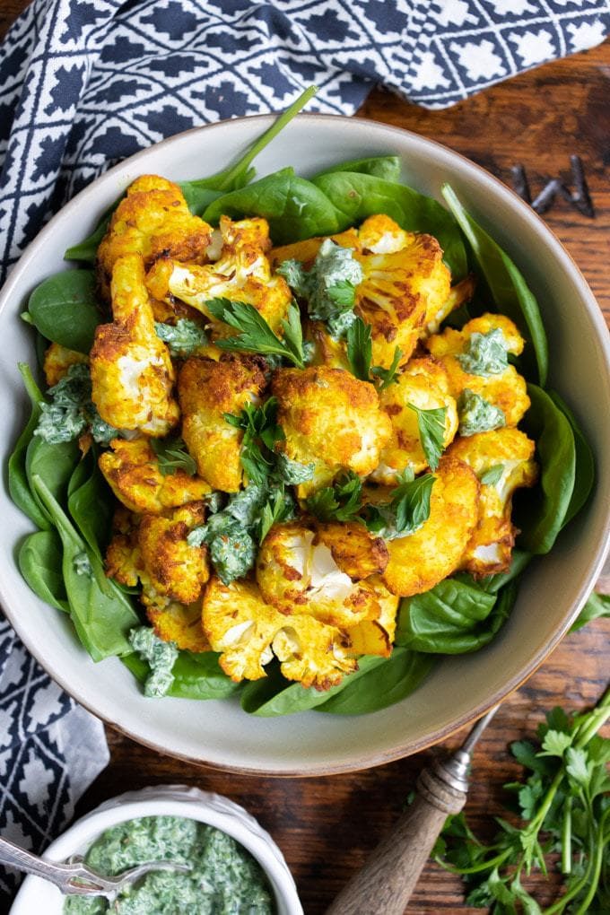 A bowl of bright yellow turmeric roasted cauliflower on a bed of spinach topped with a fresh herb dressing. 