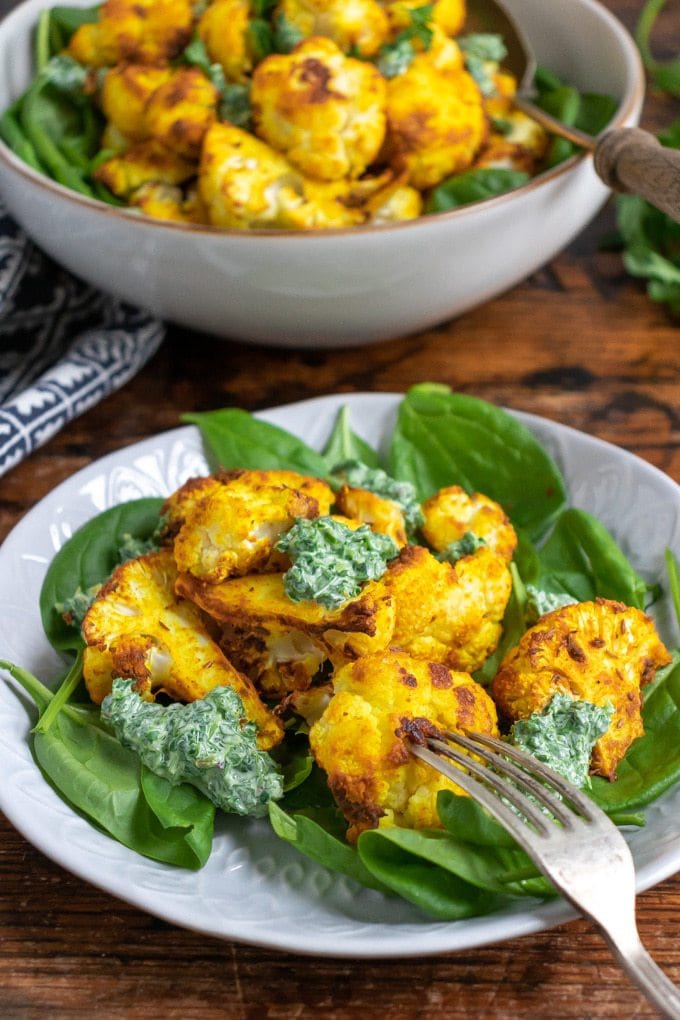 Close up of a plate with spinach and turmeric roasted cauliflower with fresh herb dressing. A white bowl of roasted cauliflower is in the background.