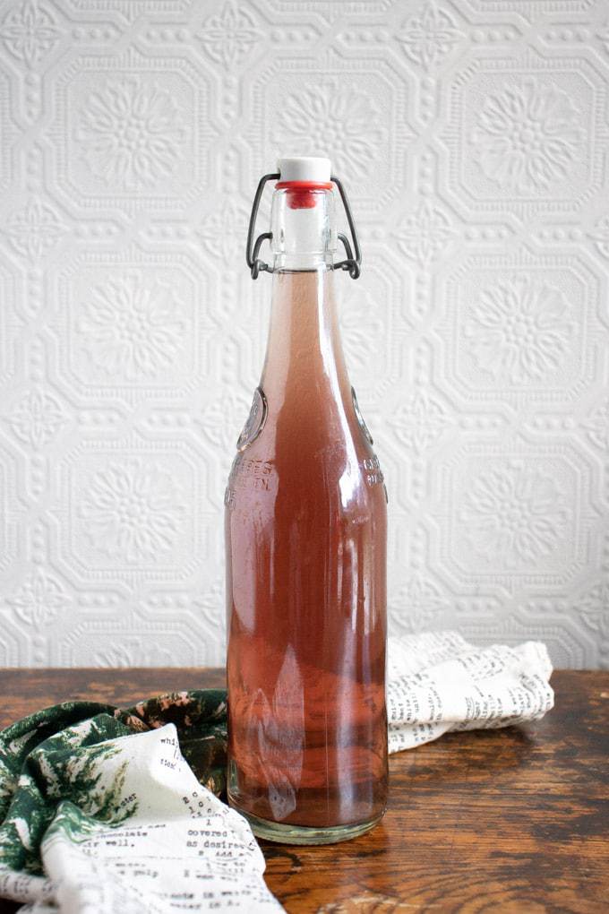 Bottle of kombucha with a green and white tea towel on a wooden table.