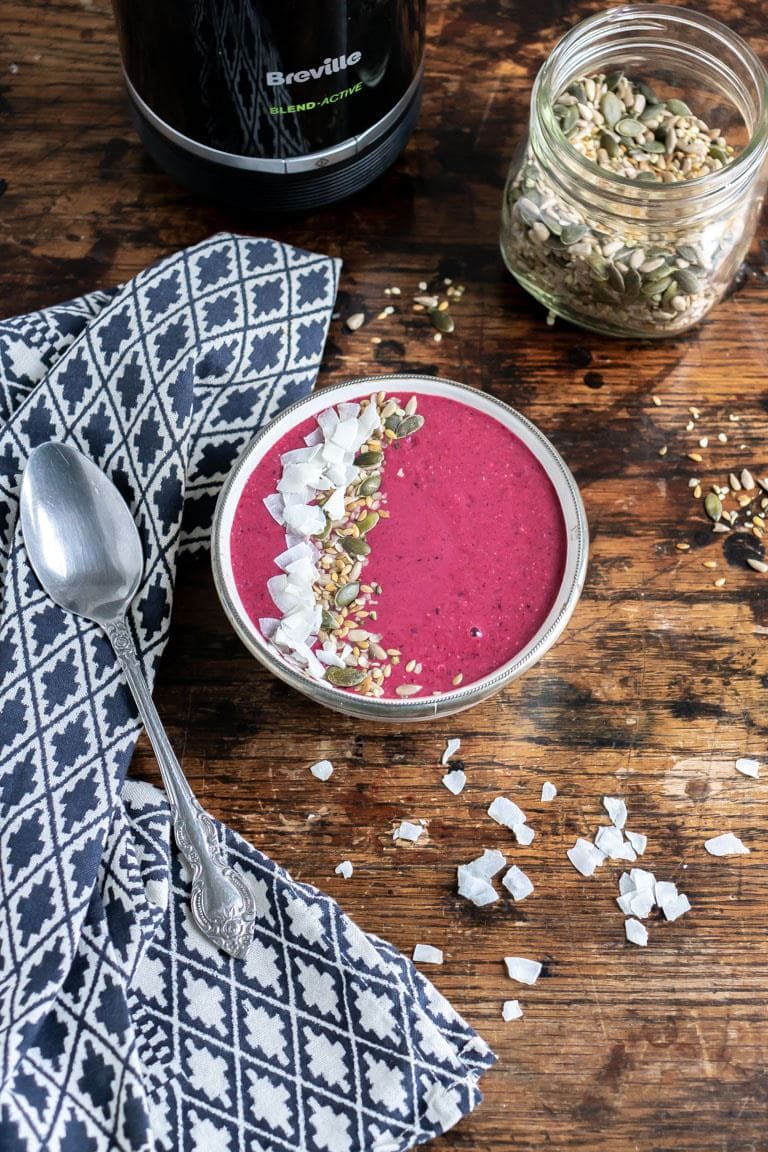 A bowl on a wooden table filled with acai smoothie, topped with seeds and coconut. Next to a blue tea towel. Get the recipe.