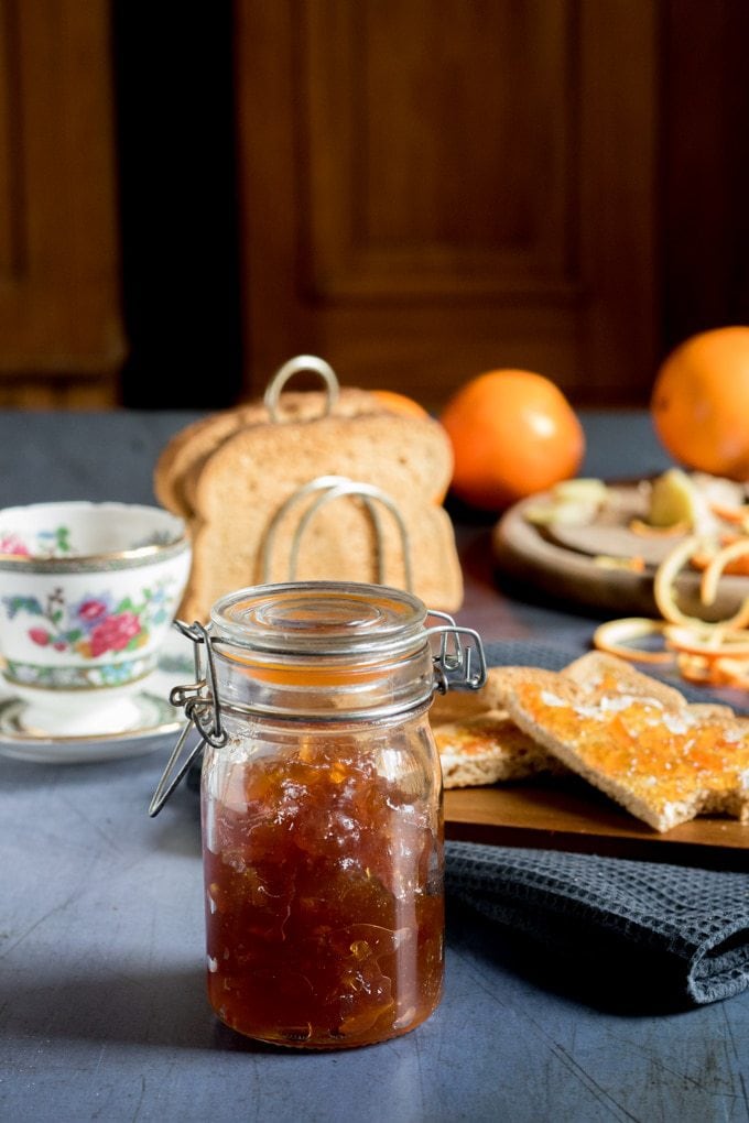 A jar of homemade ginger orange marmalade preserves recipe, in front of toast and teacups. 