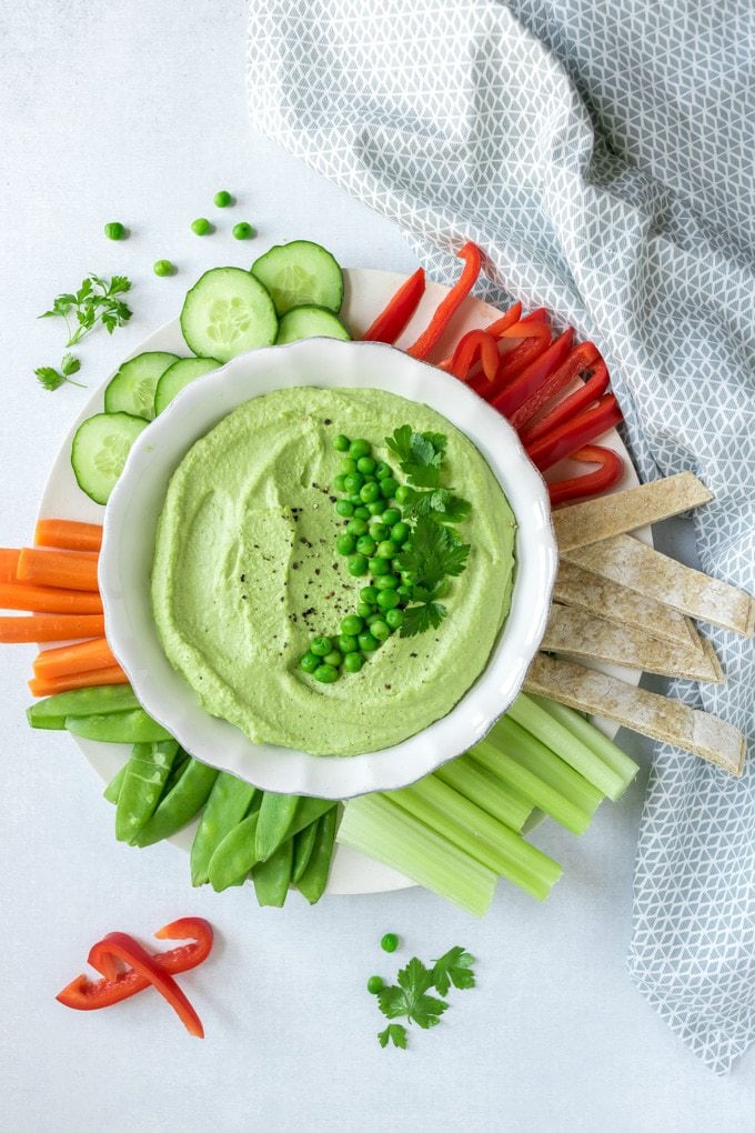 A bowl of fluffy pea hummus surrounded by raw vegetables and topped with peas and herbs. This recipe is so easy and makes a beautiful green dip. 