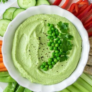 A bowl of fluffy pea hummus surrounded by raw vegetables and topped with peas and herbs. This recipe is so easy and makes a beautiful green dip.