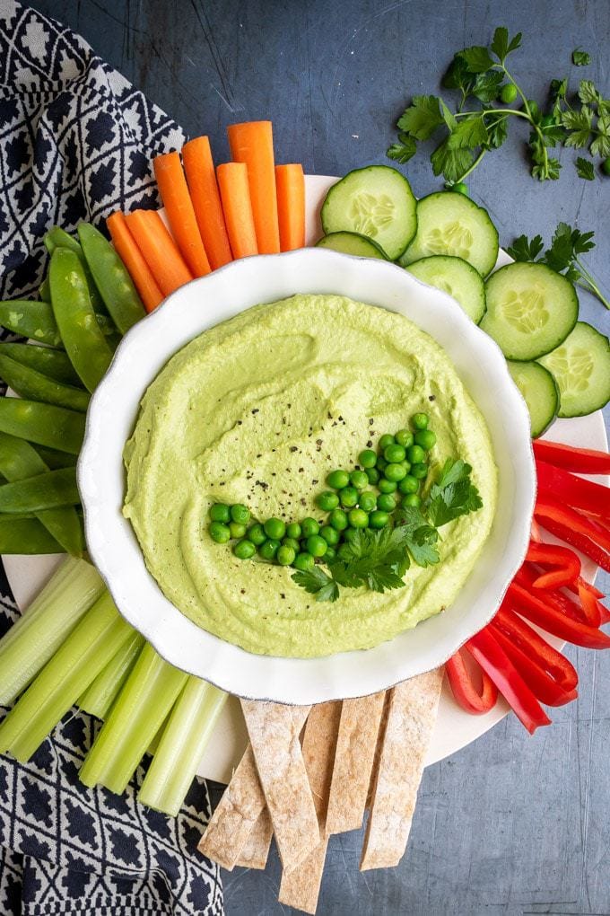 A bowl of fluffy pea hummus surrounded by raw vegetables and topped with peas and herbs. This recipe is so easy and makes a beautiful green dip. 