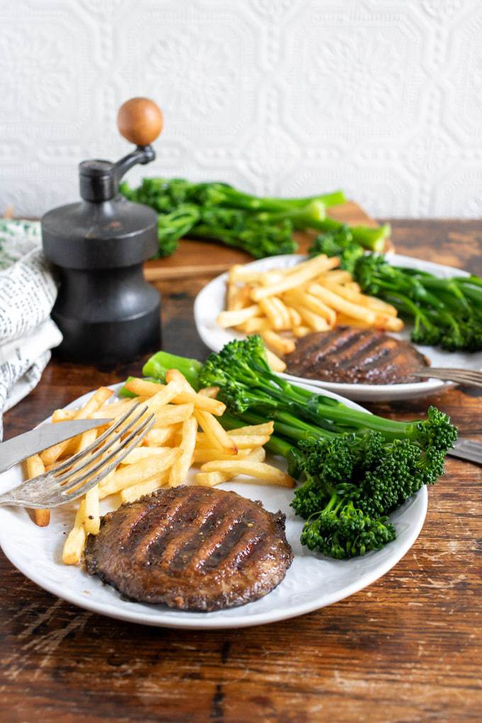 Two plates with marinated grilled mushroom steaks, served with broccolini and fries. Table set for two with this vegan recipe.