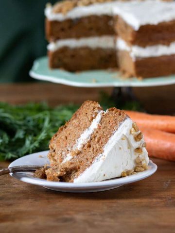 A slice of two layer vegan carrot cake. It's moist, fluffy and delicious one bowl easy recipe. Shown with vegan cream cheese frosting and walnuts.