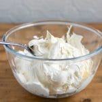 A bowl filled with thick and fluffy vegan cream cheese frosting recipe. In a glass bowl on a wooden table.