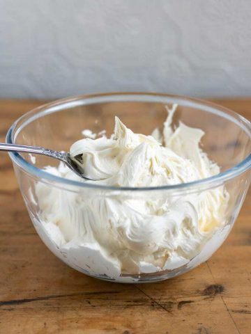 A bowl filled with thick and fluffy vegan cream cheese frosting recipe. In a glass bowl on a wooden table.