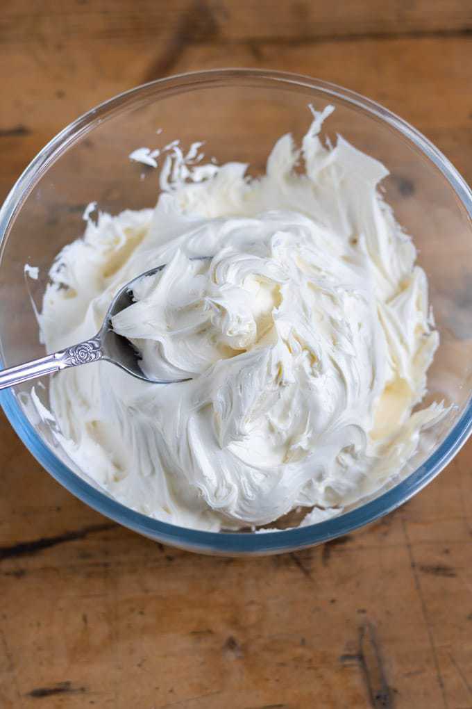 A bowl filled with thick and fluffy vegan cream cheese frosting recipe. With a spoon full of the icing. In a glass bowl on a wooden table.