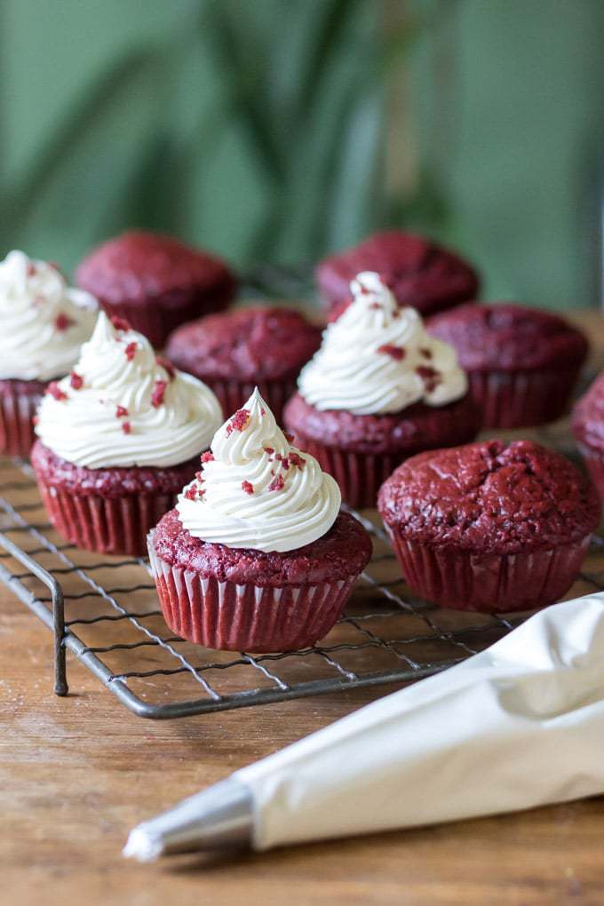 A rack of vegan red velvet cupcakes with vegan cream cheese frosting and a piping bag of frosting. Get the easy one bowl recipe. 