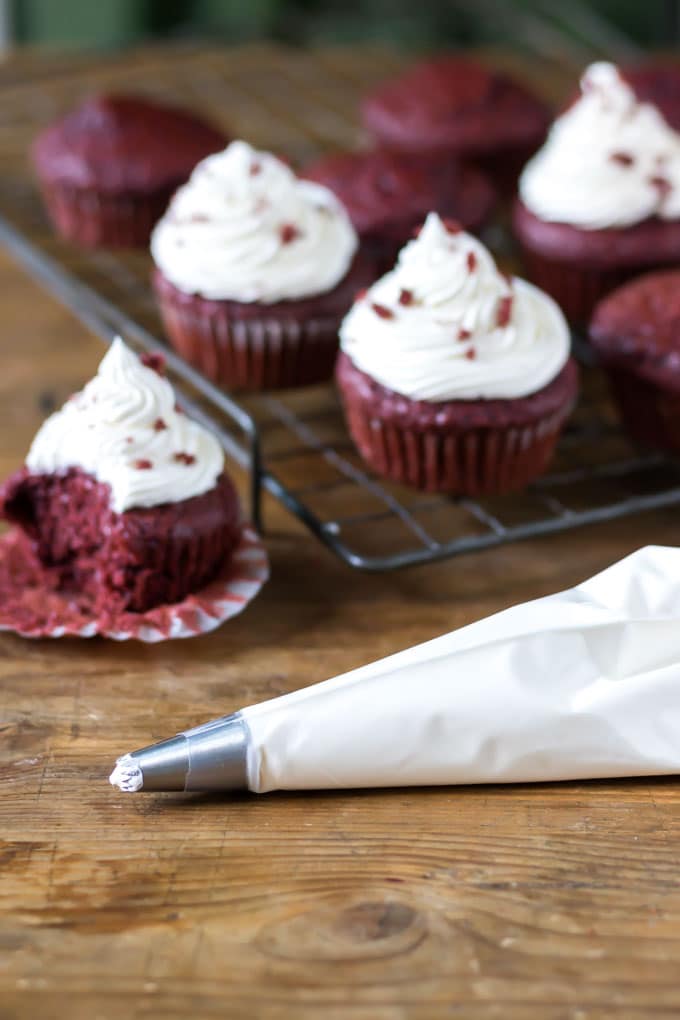 Thick creamy vegan cream cheese in a piping bag in front of frosted vegan red velvet cupcakes. 