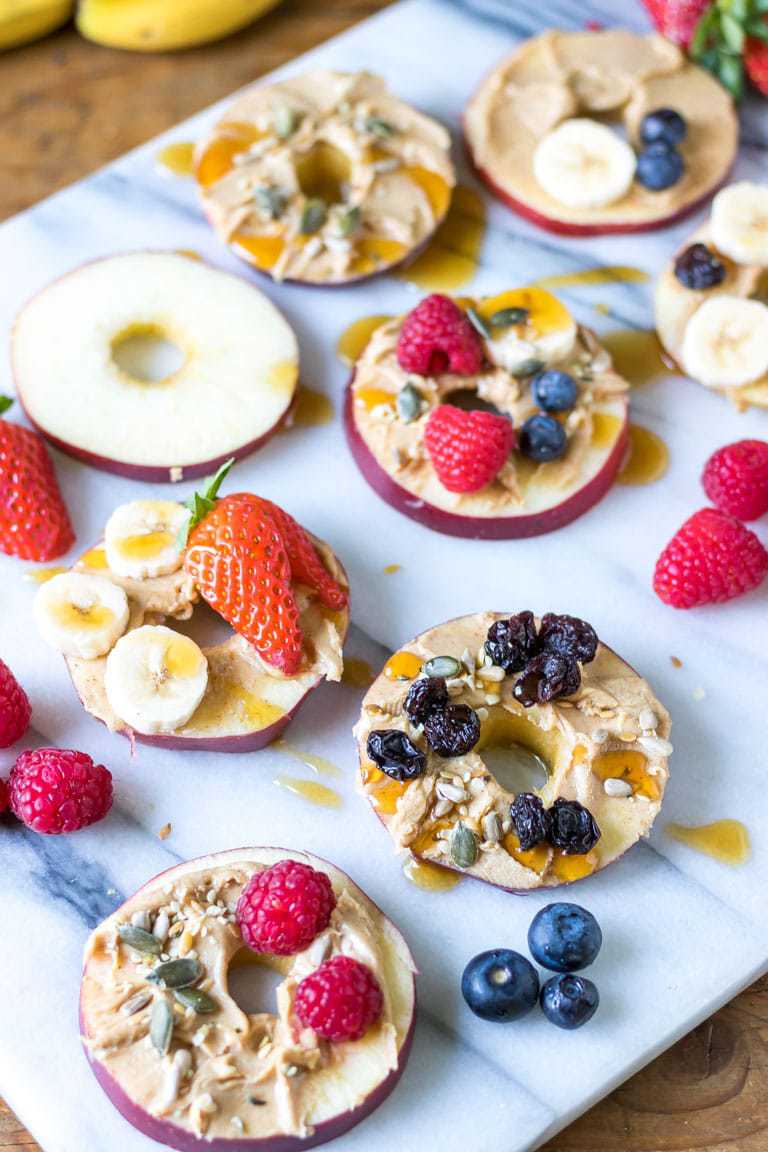 Slices of cored apple spread with peanut butter and topped with fruit, seeds and maple syrup. Vegan kids snack recipe.