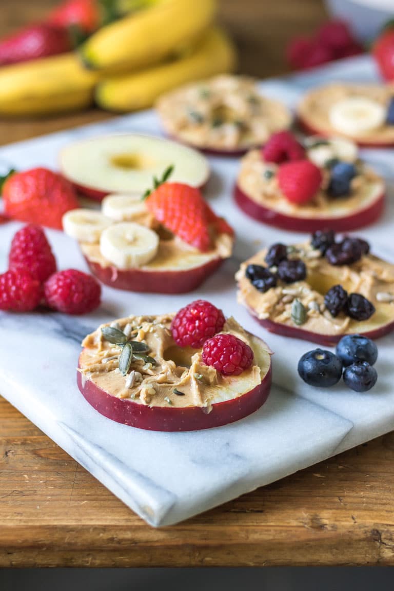 Round slices of cored apple are shown on a marble board spread with peanut butter and topped with fruit and seeds. Get the easy snack recipe now.