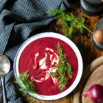 A bowl of creamy beetroot soup on a wooden table with sprigs of fresh dill.