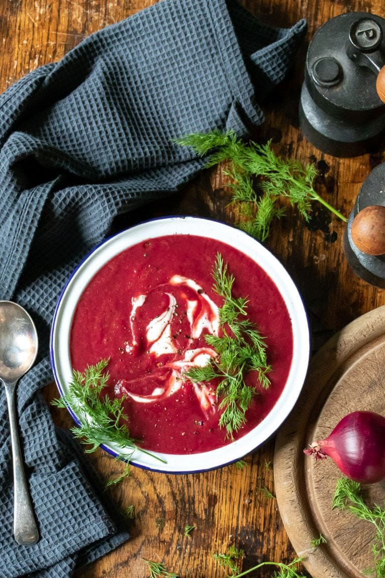 A bowl of creamy beetroot soup on a wooden table with sprigs of fresh dill. 