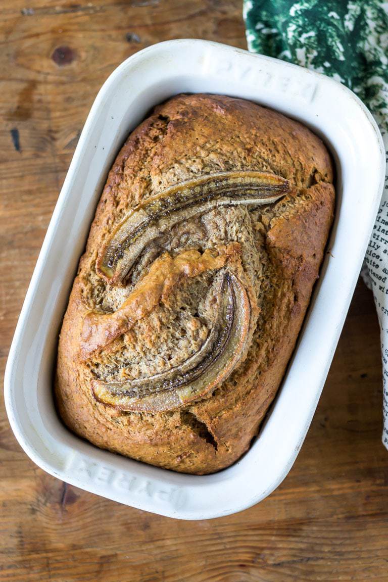 A loaf pan with freshly baked vegan banana bread with bananas decorating the top. Get the easy one bowl 10 ingredient recipe now.