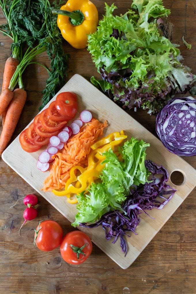 A table with vegetables sliced for a sandwich, including tomato, radishes, carrot, yellow bell pepper, lettuce, cabbage and more.