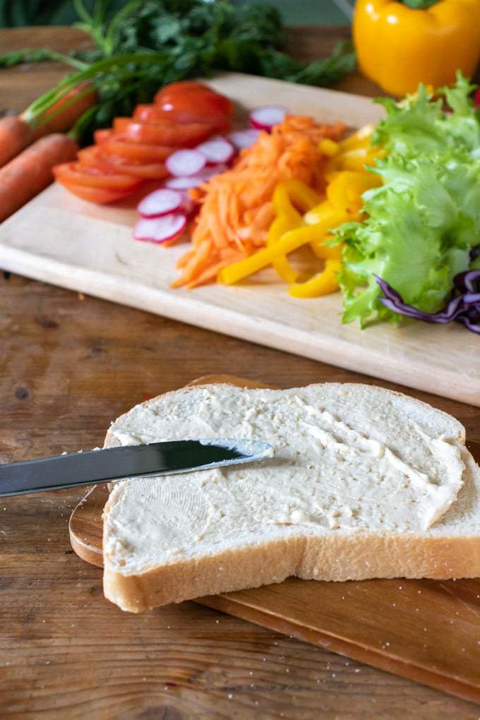 How to make a vegetable sandwich - spreading the bread with hummus in front of a tray of sliced veggies.