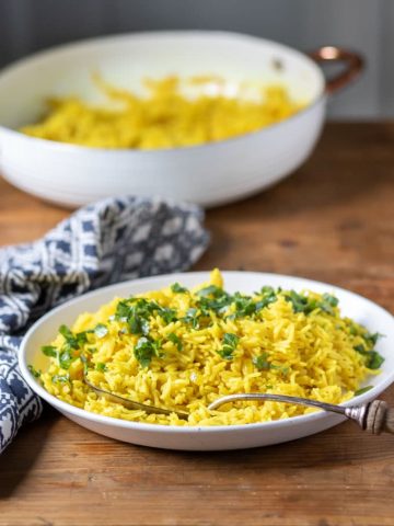 A serving bowl of yellow rice (turmeric rice recipe) topped with fresh herbs, shown with a vintage serving spoon and blue napkin.