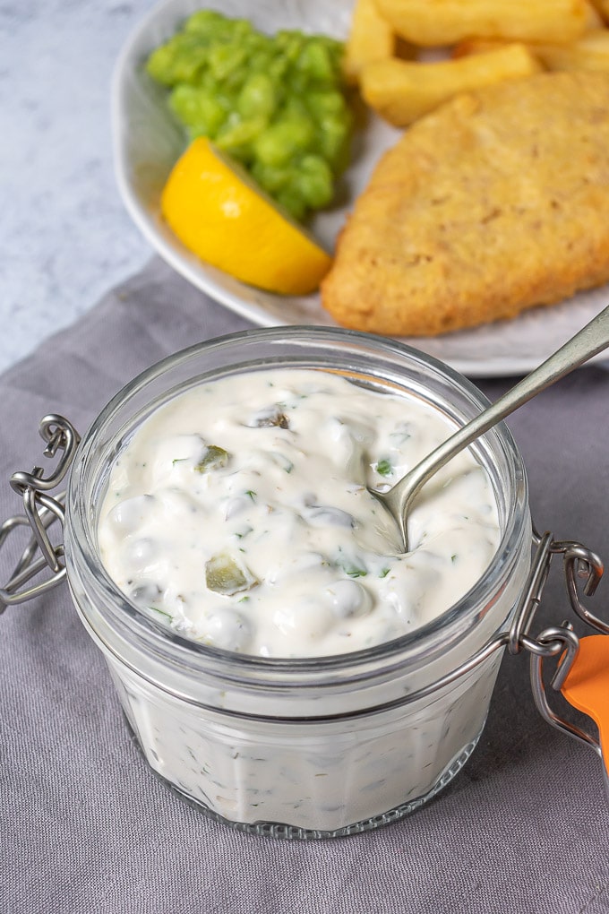 A jar of homemade tartar sauce in front of a plate of fish n chips.