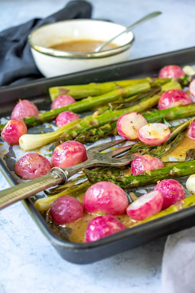 A roasted vegetable sheet pan meal with roasted radishes and asparagus with a bowl of maple mustard dressing. 