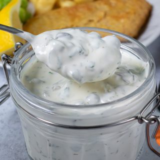 A spoonful of homemade tartare sauce coming out of a jar, next to a plate of fish and chips.