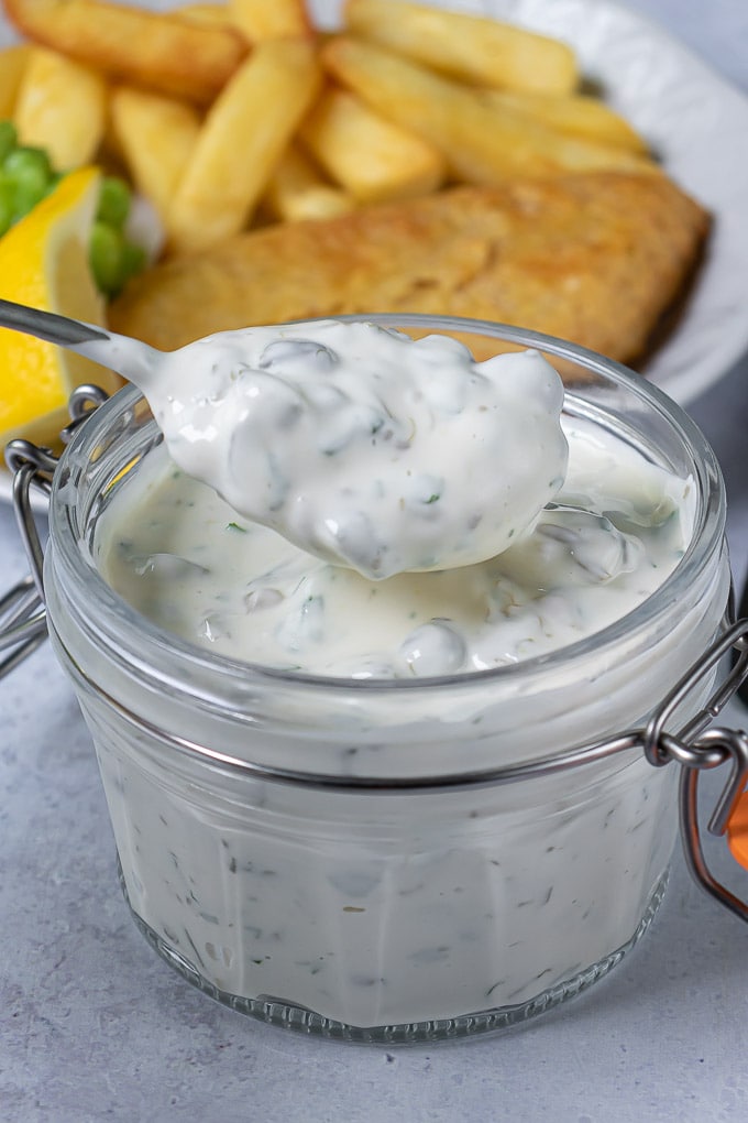 A spoonful of homemade tartare sauce coming out of a jar, next to a plate of fish and chips.