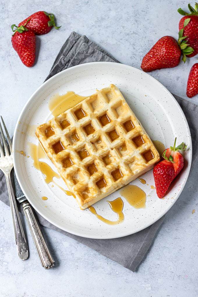 A white plate with eggless waffle recipe drizzled with maple syrup and strawberries on the side.