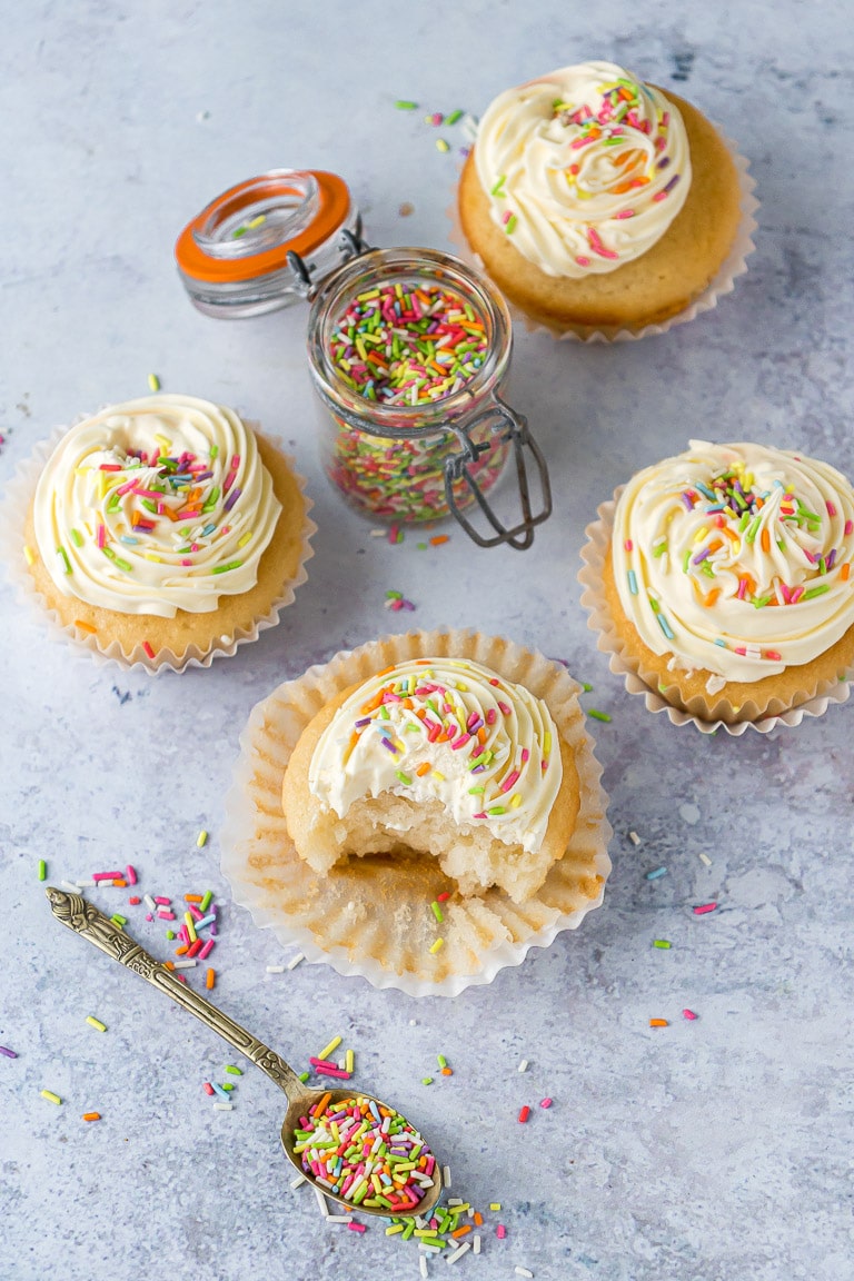 Overhead shot of vegan vanilla cupcakes with sprinkles, next to a spoonful of sprinkles.