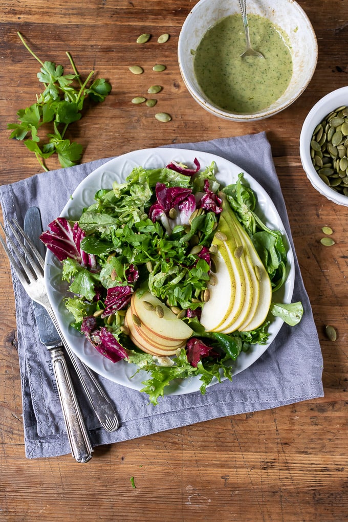 A plate of leafy green salad, with slices of apple and pear and a pumpkin seed dressing in a bowl next to it.