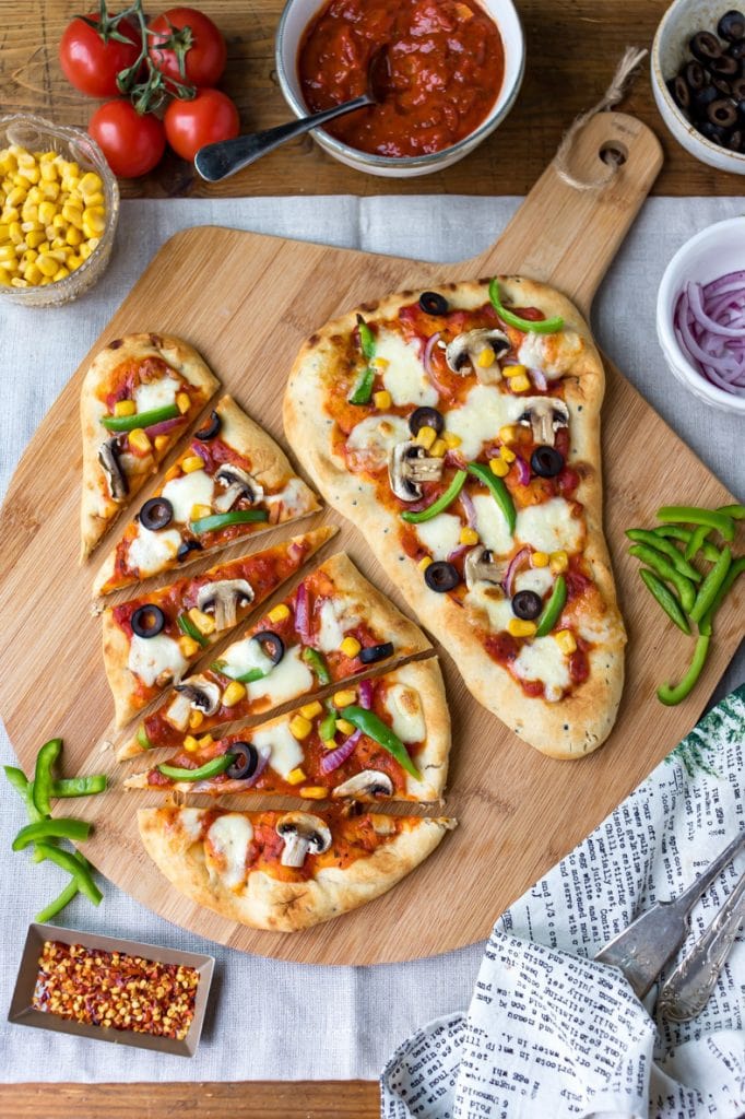 Naan bread pizza, ones sliced and the other whole, with toppings in bowls around them.