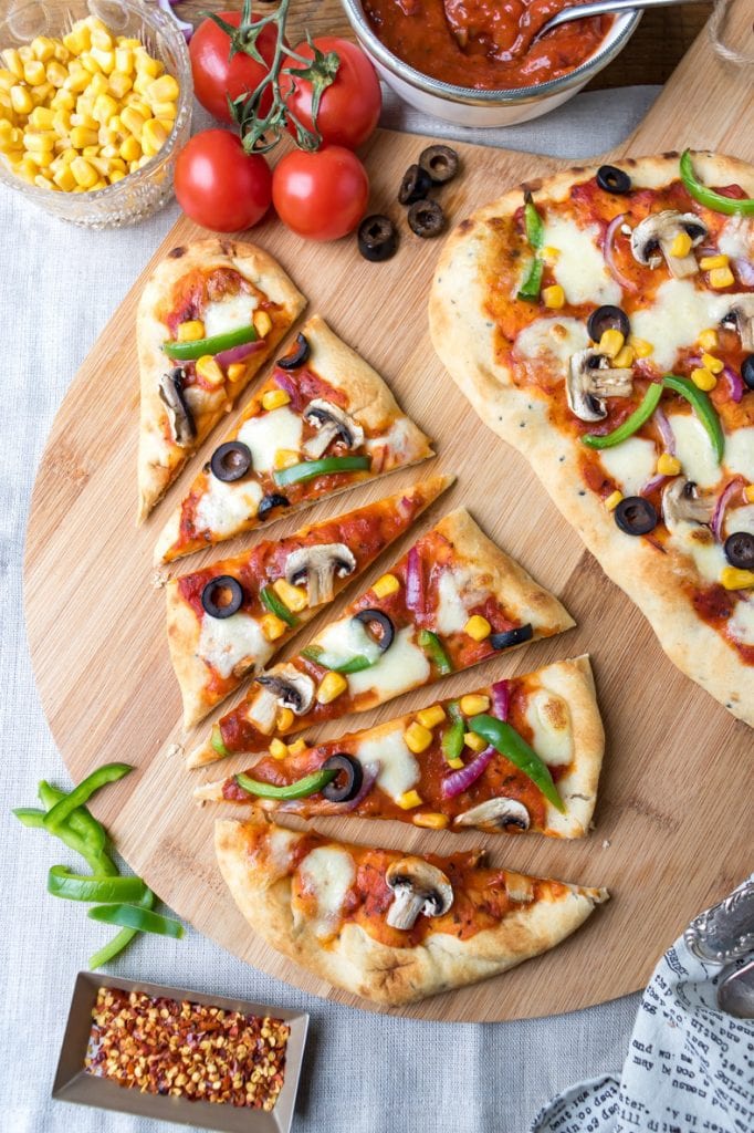 Naan Pizza, two one a wooden board, one is sliced into triangles. Toppings in dishes around them.