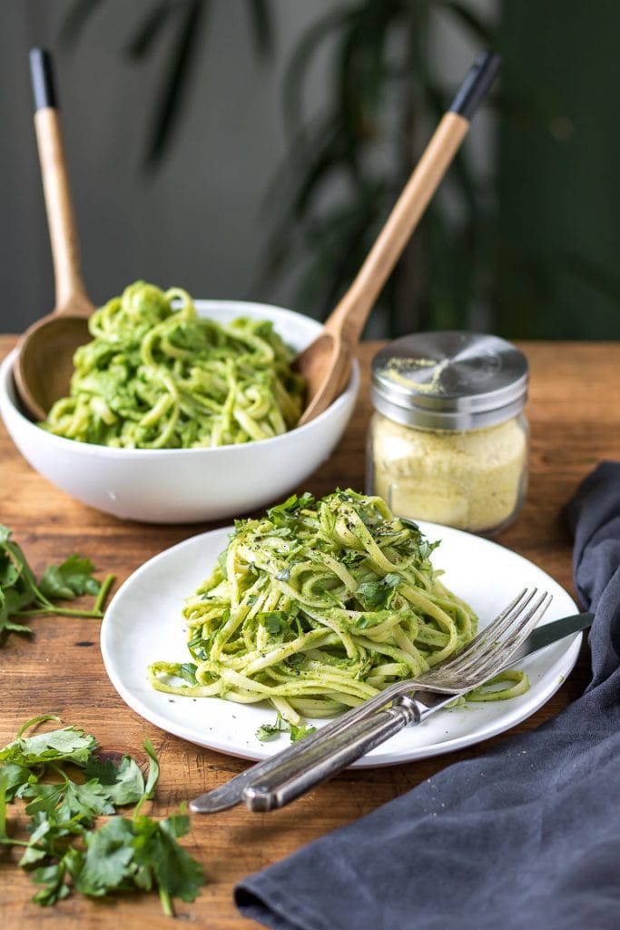 A plate of green pasta recipe in front of a serving bowl of spaghetti tossed in avocado herb sauce