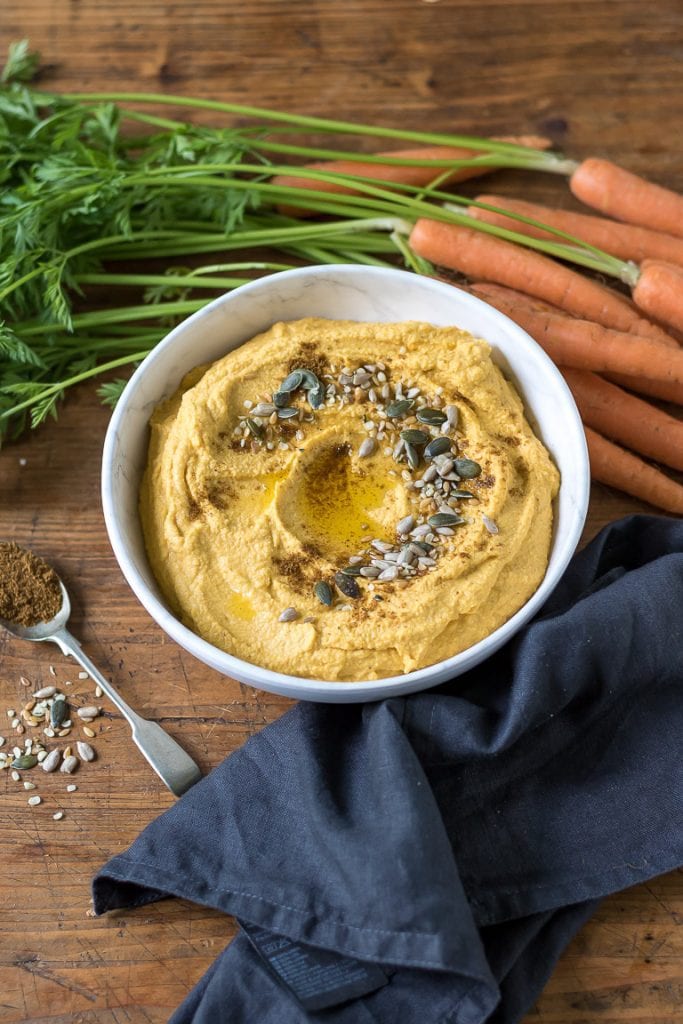 Looking down at a bowl of carrot hummus next to fresh carrots and a teaspoon of seeds.
