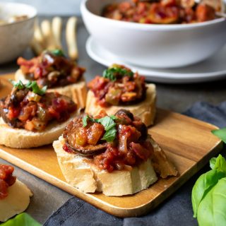 A wooden tray with Caponata alla Siciliana on rounds of toast served as an appetizer. With a bowl of the Italian eggplant stew in the background.