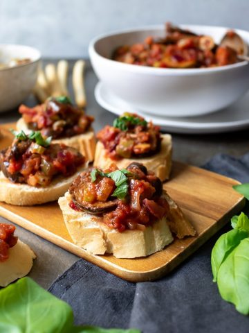 A wooden tray with Caponata alla Siciliana on rounds of toast served as an appetizer. With a bowl of the Italian eggplant stew in the background.
