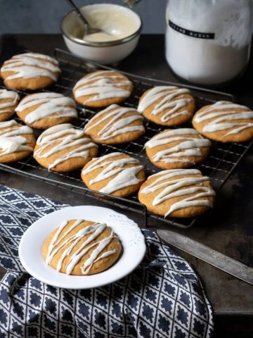Iced pumpkin cookies on a cooling rack, with a soft pumpkin cookie in front on a plate. Get the recipe now.
