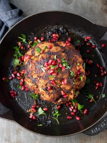 Overhead photo of a whole baked cauliflower in a skillet with pomegrante arils and fresh herbs.