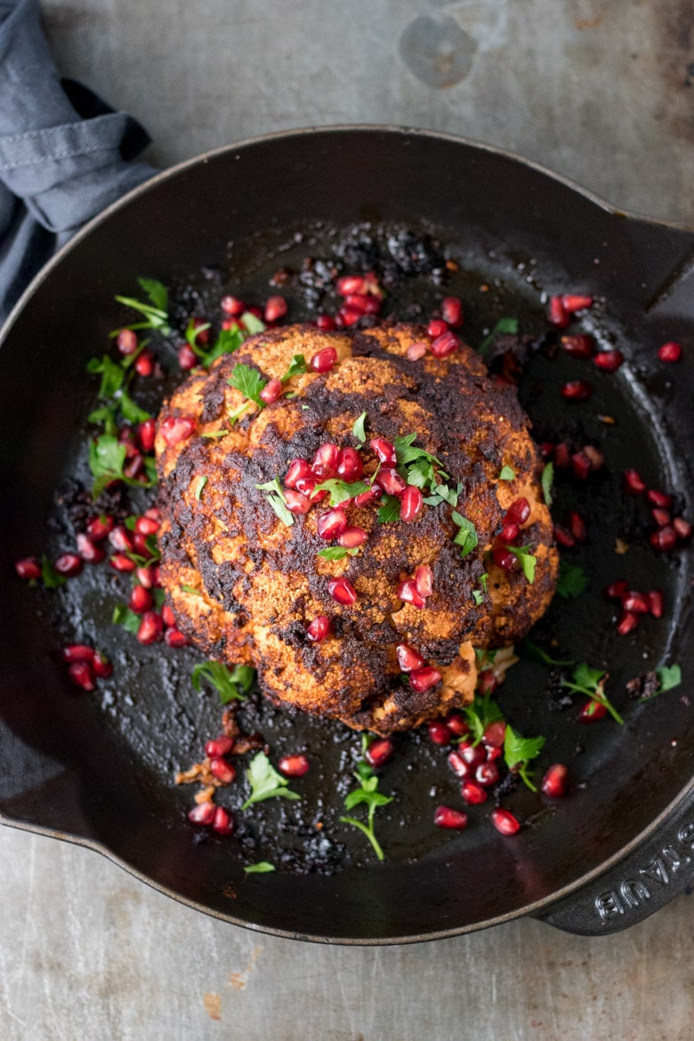 Overhead photo of a whole baked cauliflower in a skillet with pomegrante arils and fresh herbs.