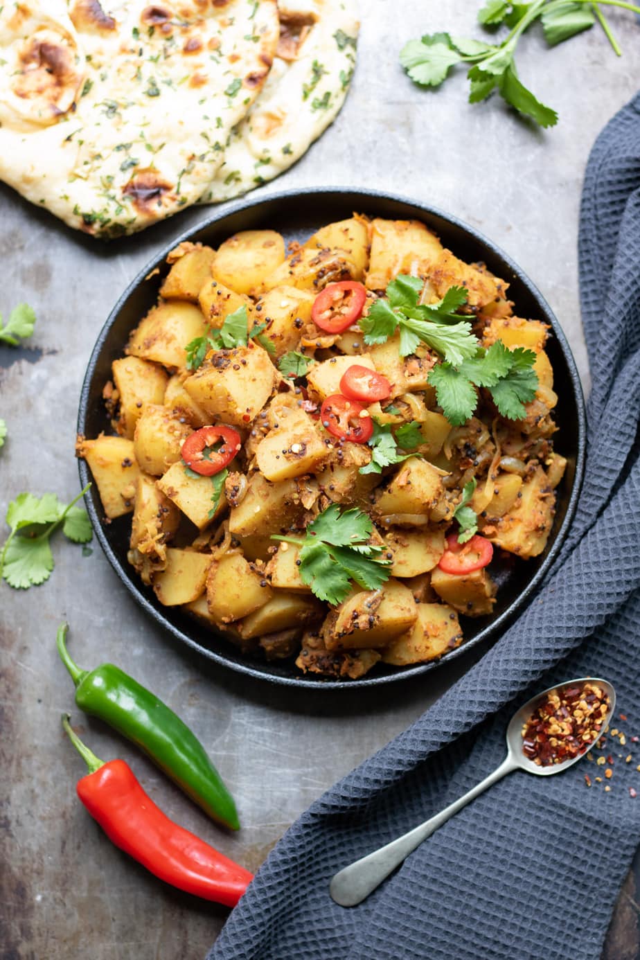 A bowl of Bombay Aloo with chillis, chilli flakes and naan bread.