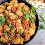 Close up of a dish of Bombay Potatoes - an Indian potato curry side dish. With naan and a teaspoon of chilli flakes.