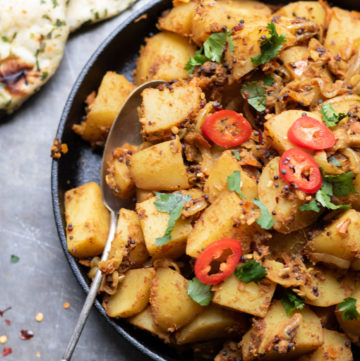 A dish of Bombay Potatoes (aka Bombay aloo Indian potato side dish), with naan and sprinkled with coriander and red chillis.