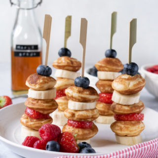 Small pancakes on sticks with sliced bananas, strawberries and blueberries with a bottle of maple syrup in the background.