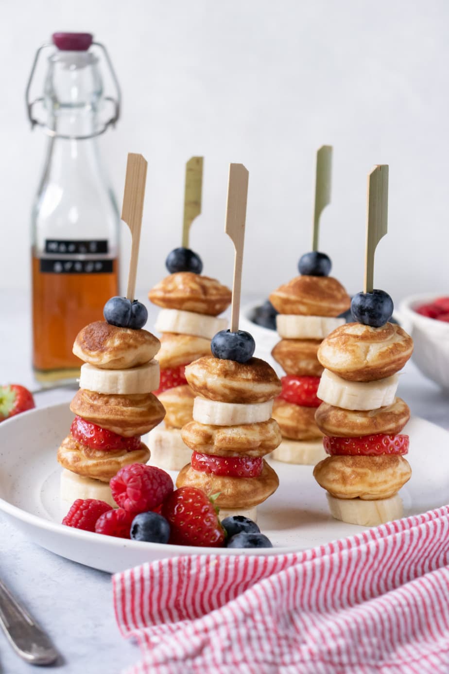 Small pancakes on sticks with sliced bananas, strawberries and blueberries with a bottle of maple syrup in the background.