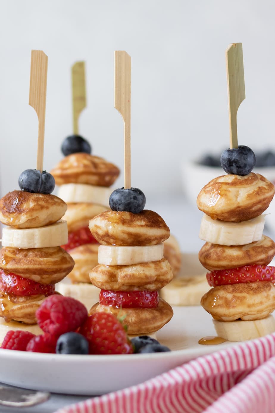 Close up of pancakes on sticks with bananas, strawberries and blueberries, drizzled with maple syrup.