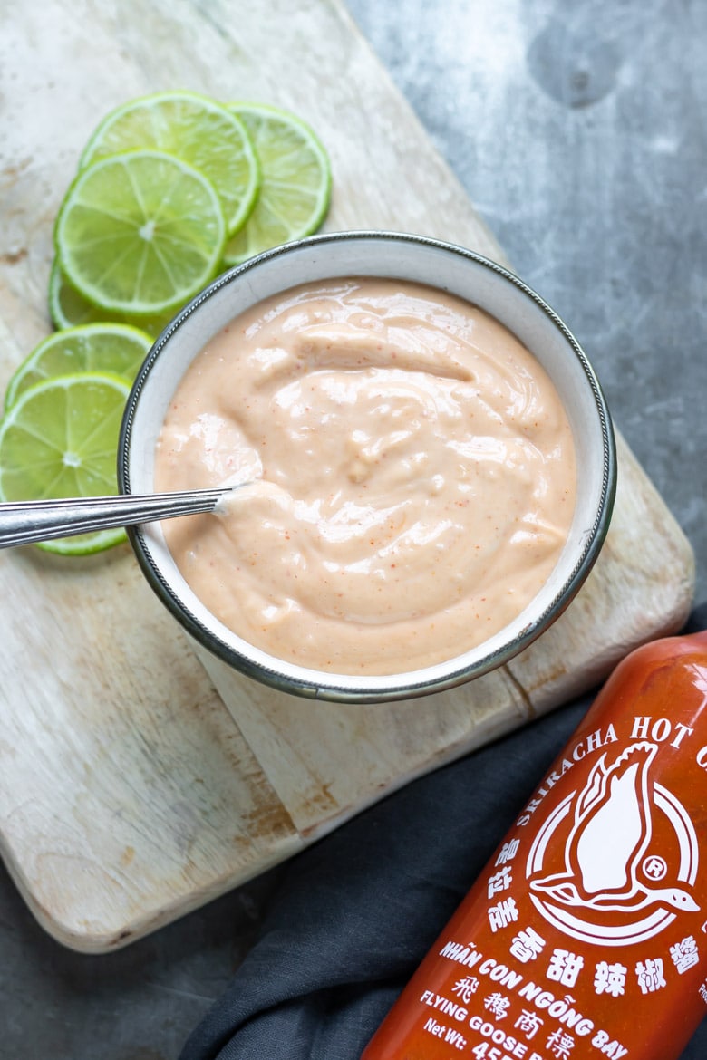 A bowl of sriracha aioli next to slices of lime and a bottle of sriracha hot sauce.