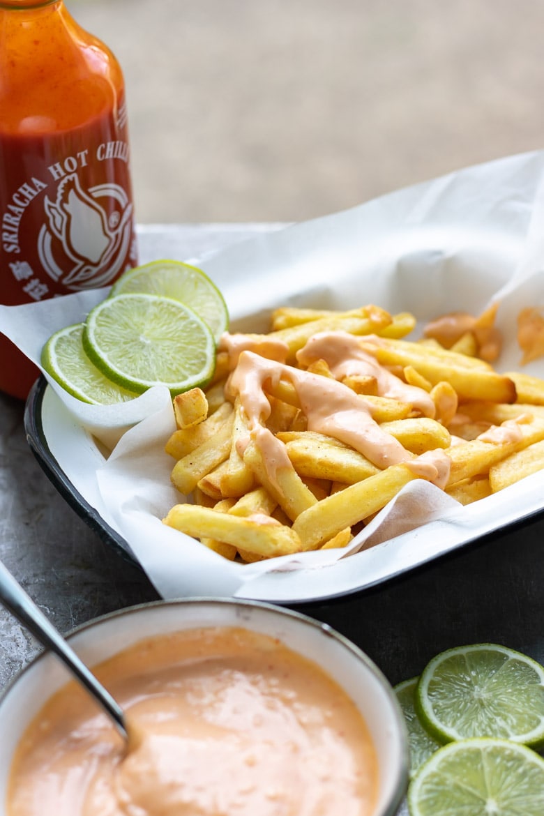 A bowl of sriracha aioli next to slices of lime and a bottle of sriracha hot sauce.