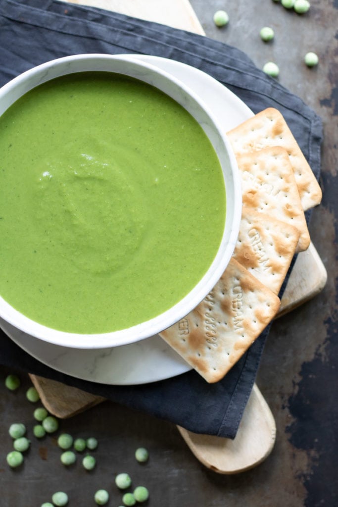A bowl of thick creamy pea soup next to frozen peas and crackers.