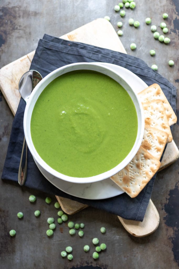 A bowl of thick creamy pea soup next to frozen peas and crackers